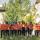 Des personnes portant des t-shirts orange debout devant une rangée d'arbres. À l'avant-plan, on voit des herbes hautes. Sur les t-shirts, de petites empreintes de main sont regroupées en forme de cœur avec le numéro 215+ au-dessus.